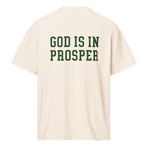 Puede incluir: Camiseta color crema con la frase "GOD IS IN PROSPER" impresa en letras verdes oscuras en la espalda. La camiseta tiene un corte holgado y mangas cortas, adecuada para uso casual.
