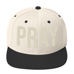May include: A cream-colored flat-brimmed baseball cap with the word "PRAY" in a textured, light beige font. The brim is black, and the cap has a black button on top.