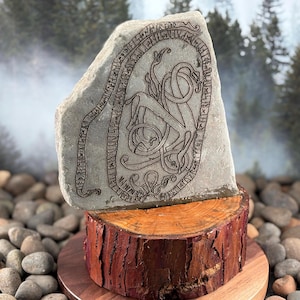 May include: A gray stone carving featuring a detailed engraved design of a serpent and other symbols. The stone is mounted on a wooden base, showcasing the artwork. The background includes a blurred forest scene.