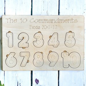 May include: A wooden puzzle with the numbers 1 to 10 cut out. The text "The 10 Commandments Exodus 20:1-17" is engraved at the top. The puzzle is made of light-coloured wood, ideal for educational play.