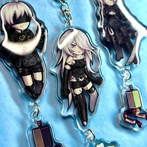 May include: A collection of acrylic keyrings featuring anime-style characters with silver hair and black clothing. The keyrings are attached to silver metal rings. The background is a light blue colour.