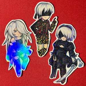 以下が含まれることがあります： ビデオゲーム「ニーア オートマタ」のキャラクターが描かれたホログラフィックステッカー3枚。 ステッカーは赤い背景に貼られています。 キャラクターは、白い髪と黒いドレスを着た若い女性、白い髪と白黒の衣装を着た若い男性、白い髪と白黒の衣装を着て剣を持った若い女性の3人です。