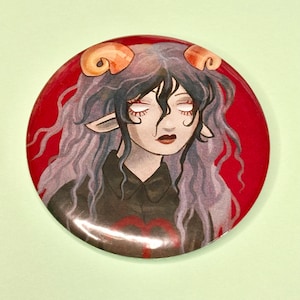 May include: A circular pin-back button featuring an illustration of a character with long wavy purple hair, orange ram horns, and pointed ears. The character wears a black collared shirt against a solid red background with a red zodiac symbol.