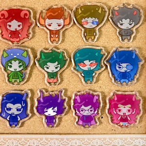 Könnte beinhalten: Eine Sammlung von zwölf Acryl-Charms mit Cartoon-Charakteren, jedes mit einem einzigartigen Design und Farbschema. Die Charaktere sind in drei Reihen zu je vier Stück angeordnet, mit einem transparenten Rand um jeden Charm. Der Hintergrund ist eine hellbraune Pinnwand.