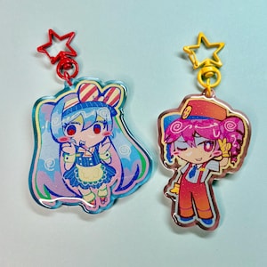 May include: Two anime-style acrylic charms. One features a blue-haired character with a red star clasp. The other charm has a pink-haired character with a yellow star clasp. Both charms have colorful designs and are likely keychains.