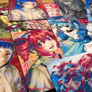 May include: A collection of colorful anime-style art prints featuring various characters with vibrant hair and clothing. The prints showcase a range of expressions and poses, with a focus on detailed facial features and dynamic compositions. Some prints include text.
