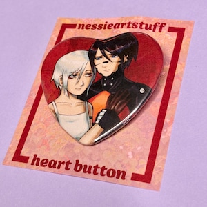 Puede incluir: Botón en forma de corazón con una ilustración de estilo anime de dos personajes. El botón tiene un fondo rojo y el texto "heart button". El botón está sobre una tarjeta rosa con el texto "nessieartstuff".