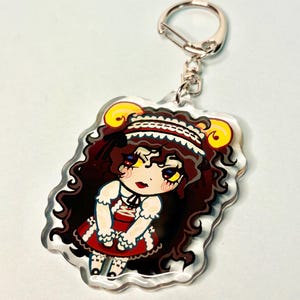 Puede incluir: Un llavero acrílico transparente con una ilustración de dibujos animados de un personaje con cabello rizado castaño oscuro, un vestido rojo y cuernos amarillos. El llavero tiene un cierre plateado.