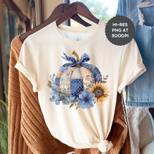 May include: A cream-coloured t-shirt with a watercolour design of a patchwork pumpkin with a blue bow, surrounded by blue flowers and a sunflower. The t-shirt is hanging on a wooden hanger. Text on the shirt reads "HI-RES PNG AT 300DPI".