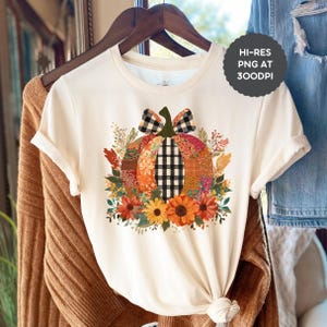 Könnte beinhalten: Cremefarbenes T-Shirt mit einem farbenfrohen Kürbisdesign. Der Kürbis besteht aus verschiedenen gemusterten Stoffen mit einer schwarz-weiß karierten Schleife und einem Stiel. Sonnenblumen und Herbstlaub umgeben den Kürbis.
