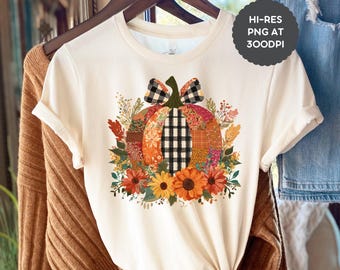Puede incluir: Camiseta color crema con un diseño de calabaza colorida. La calabaza está hecha de varias telas estampadas, con un lazo y un tallo a cuadros blancos y negros. Girasoles y follaje otoñal rodean la calabaza.