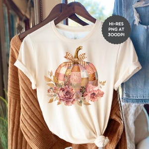 May include: Cream-colored t-shirt featuring a patchwork pumpkin design with a plaid bow and floral accents. The pumpkin is composed of various patterned fabric pieces in shades of pink, orange, and brown. The shirt is hanging on a wooden hanger.