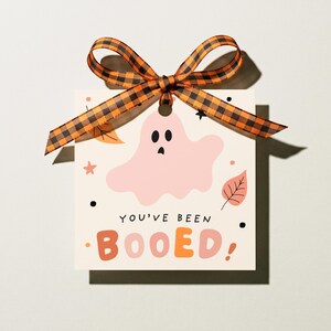 May include: A square off-white gift tag featuring a light pink ghost illustration, orange and pink autumn leaves, and the text "YOU'VE BEEN BOOED". The tag is topped with an orange and black buffalo plaid ribbon tied in a decorative bow.