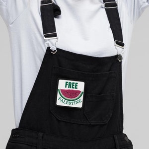 May include: Black denim overalls with adjustable straps and a front pocket featuring a patch with a watermelon design and the words "FREE PALESTINE". The person is wearing a light blue t-shirt.