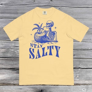 Könnte beinhalten: Ein gelbes T-Shirt mit einer blauen Grafik eines Skeletts, das aus einer Kokosnuss trinkt, mit dem Text "STAY SALTY" darunter.