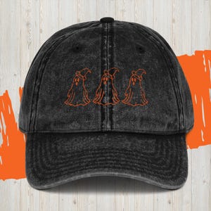 May include: A black distressed denim baseball cap with an embroidered design of three ghosts wearing witch hats. The ghosts are orange.