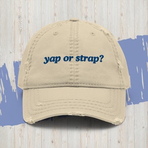 May include: A beige distressed baseball cap with the text "yap or strap?" embroidered in blue.