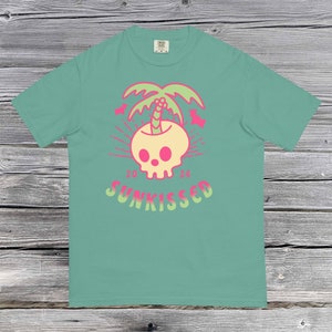 Könnte beinhalten: Ein türkisfarbenes T-Shirt mit einem Aufdruck eines gelben Schädels, aus dem ein rosafarbener Palmenbaum wächst. Der Text "20 24 SUNKISSED" ist unter dem Schädel aufgedruckt.