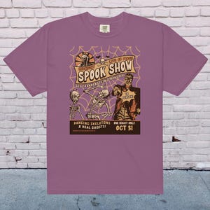 May include: Purple t-shirt featuring a vintage-style illustration of a spooky show with dancing skeletons, a monster, and the text "MIDNIGHT LIVE ON STAGE SPOOK SHOW SEE FRANKENSTEINS MONSTER IN PERSON DANCING SKELETONS & REAL GHOSTS! ONE NIGHT ONLY OCT 31".