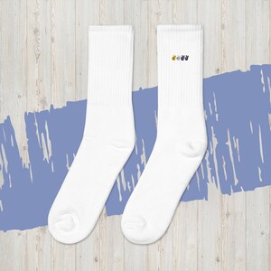 Puede incluir: Un par de calcetines blancos acanalados con un pequeño diseño bordado en amarillo, verde y morado en la parte superior. Los calcetines se muestran sobre una superficie de madera clara con un diseño pintado de azul.