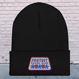 May include: Black beanie with a white patch that says "PROTECT TRANS KIDS" in blue and red letters with three pink and blue stars.
