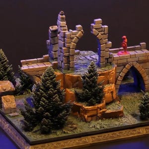 Op de afbeelding: Miniatuur fantasie diorama met een stenen brug en boog, bekroond door een klein rood figuur. De scène bevat miniatuurbomen en grasachtig terrein. De tekst "DUNGEON BLOCKS XL" is zichtbaar.