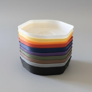 Puede incluir: Una pila de bandejas hexagonales multicolores. Los colores incluyen blanco, amarillo, naranja, rojo, azul marino, morado, verde, marrón, gris y negro. Las bandejas están apiladas, mostrando sus diferentes tonos.