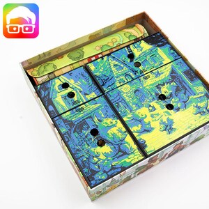May include: A rectangular board game box containing four square cardboard tiles with a vibrant blue and yellow illustration of dragons and stone buildings. The tiles feature circular cutouts and are arranged inside the box alongside a game board.