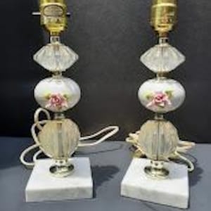 May include: Pair of vintage table lamps with a marble base. Each lamp features a gold-toned socket, clear faceted glass, a white ceramic section with pink rose details, and a clear glass base. The lamps are connected by white cords.