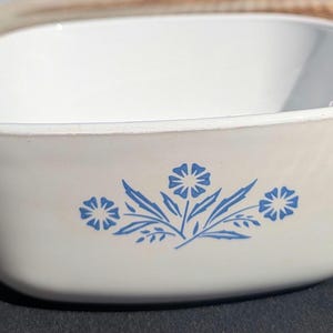 May include: A white square casserole dish with a blue floral design. The dish has two handles and is made of a durable material. The floral pattern includes blue flowers and leaves. This dish is suitable for baking and serving.