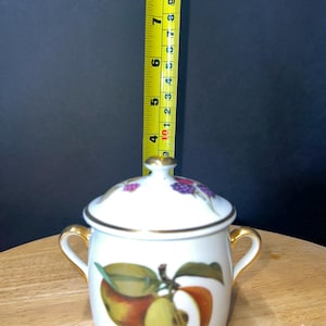 May include: White ceramic jar with gold trim and two handles. The jar has a lid with a floral design and is decorated with a painting of apples. The jar is approximately 4 inches tall.