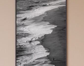 May include: A black and white photograph of a beach with waves crashing on the shore. A single person walks along the beach in the distance.