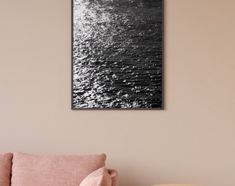 May include: A black and white photograph of rippling water with sunlight reflecting off the surface, framed in a black frame and hanging on a light brown wall.