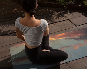 Puede incluir: Una persona sentada en una esterilla de yoga con un diseño de puesta de sol. La esterilla es azul, naranja y amarilla. La persona lleva un top corto gris y pantalones negros.