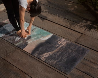 Puede incluir: Una persona haciendo yoga en una esterilla de yoga impresa con una escena de playa. La esterilla tiene una combinación de colores azul, verde y marrón y presenta una costa rocosa con olas rompiendo en la orilla.