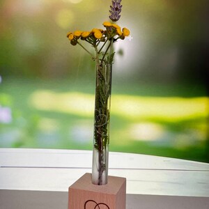 Könnte beinhalten: Ein kleiner Holzblock mit einem eingravierten Herz auf der Vorderseite. Eine Glas-Reagenzglasvase mit gelben Blumen und lila Lavendel ist in die Oberseite des Blocks gesteckt.