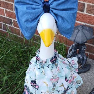 May include: A white goose statue wearing a large blue denim bow with a dark blue center knot and a light blue ruffled dress featuring a repeating pattern of hot air balloons, clouds, and birds with pink and dark blue accents on the fabric.
