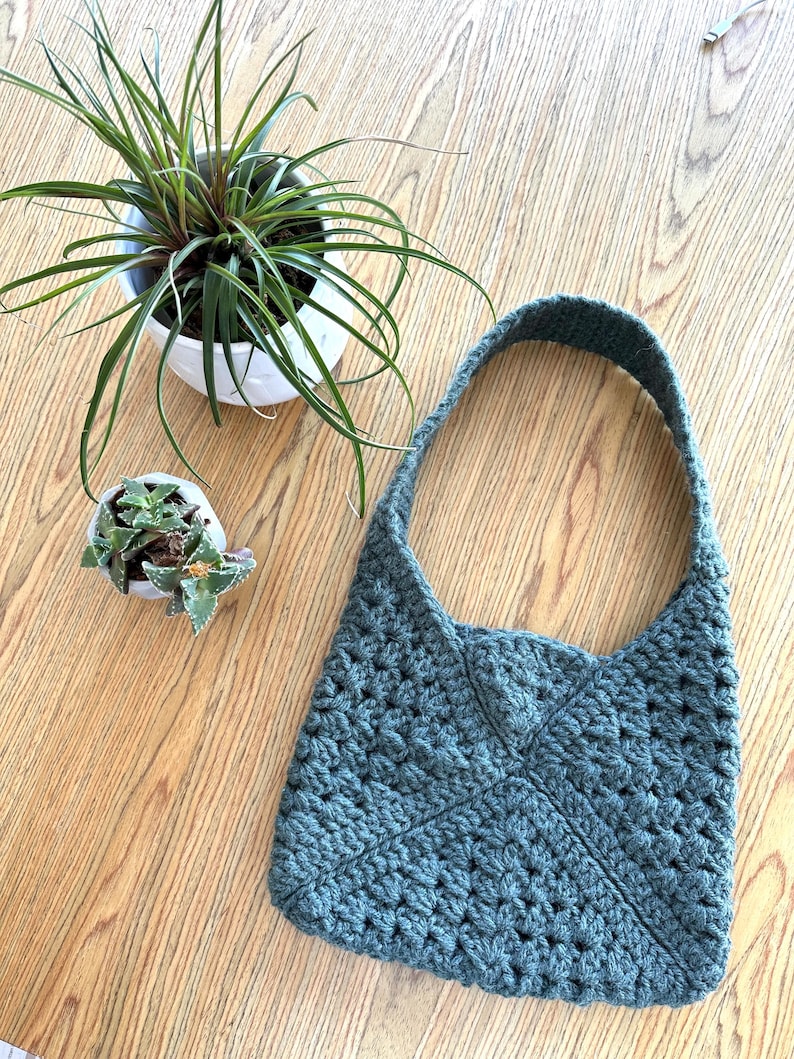 A granny square crochet bag on a table next to 2 small plants. The crochet bag is made with granny squares sewn together, the strap is also crocheted.
