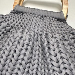 May include: A dark gray handbag featuring a thick, braided knit texture made from fabric cord. The bag is attached to a smooth, light-toned wooden handle with a rounded rectangular shape, securely integrated into the top of the woven structure.
