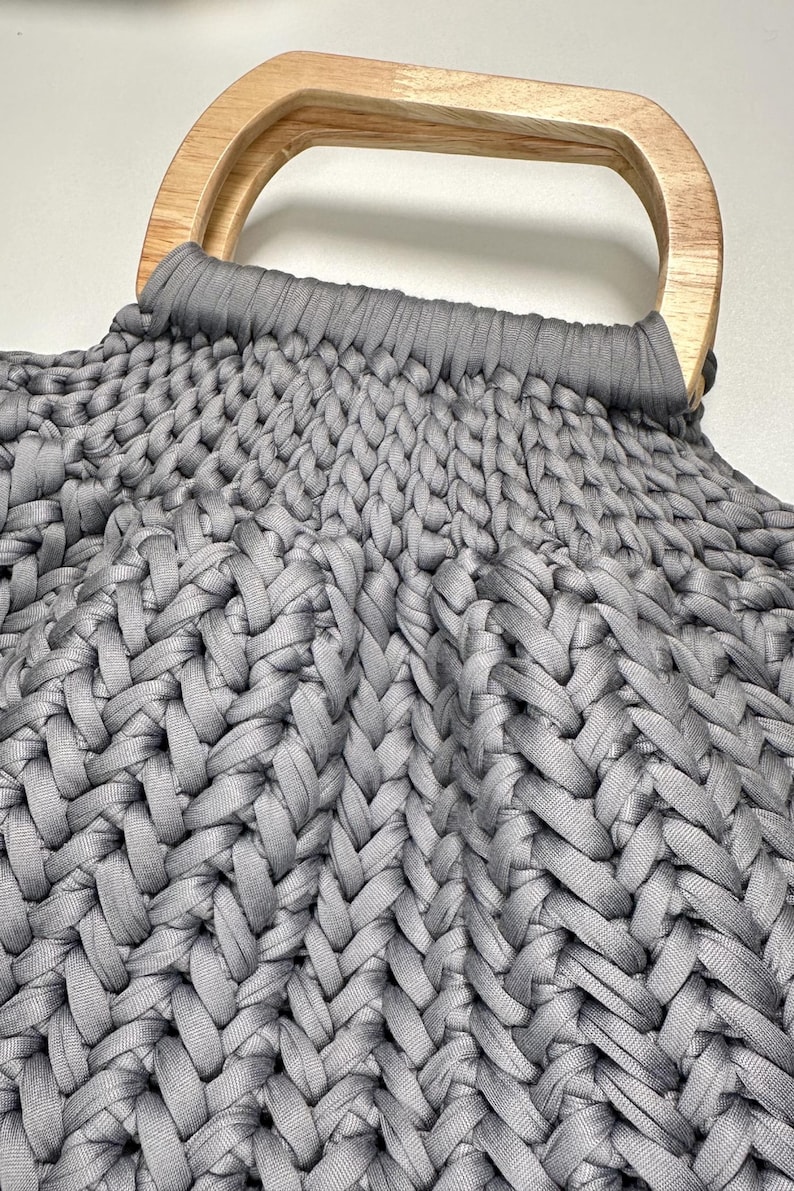 May include: A dark gray handbag featuring a thick, braided knit texture made from fabric cord. The bag is attached to a smooth, light-toned wooden handle with a rounded rectangular shape, securely integrated into the top of the woven structure.