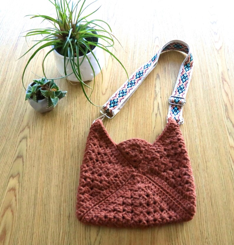 A crossbody crochet bag on a table next to 2 small plants. The crochet bag is made with granny squares sewn together. The colorful strap gives a modern look.