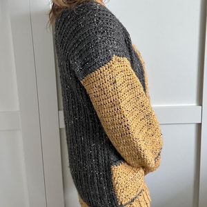 A gray and mustard yellow crochet sweater with a textured knit like design. The sleeves and bottom hem are gold, while the body is gray with flecks of white. The sweater has a relaxed fit and a round neckline.