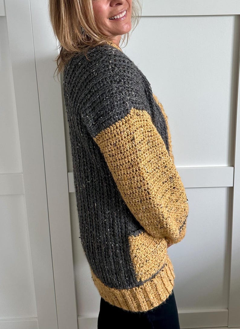 A gray and mustard yellow crochet sweater with a textured knit like design. The sleeves and bottom hem are gold, while the body is gray with flecks of white. The sweater has a relaxed fit and a round neckline.