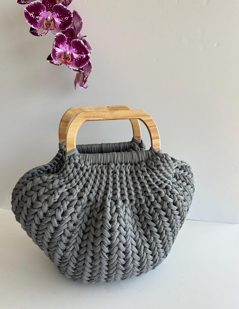 A gray, chunky crochet handbag with wooden handles. The bag has a textured, crochet herringbone stitch pattern.
