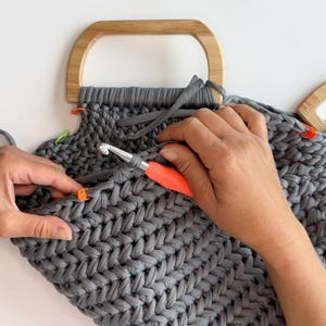 May include: A dark gray crocheted bag in progress featuring a herringbone stitch pattern, attached to a light-colored wooden D-shaped handle. The project includes a metal crochet hook with an orange handle and several small plastic stitch markers.