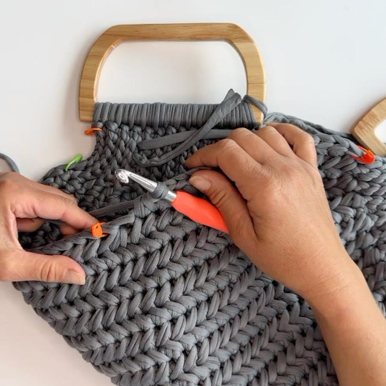 May include: A dark gray crocheted bag in progress featuring a herringbone stitch pattern, attached to a light-colored wooden D-shaped handle. The project includes a metal crochet hook with an orange handle and several small plastic stitch markers.