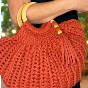 May include: A rust-colored crocheted handbag featuring a thick, braided yarn texture. The bag includes two rounded, light-colored wooden handles and a decorative tassel accent attached to the top edge with three small, round wooden beads.