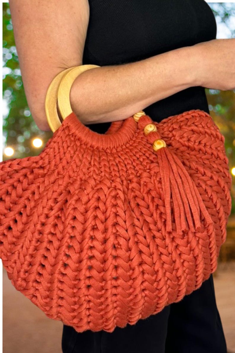 May include: A rust-colored crocheted handbag featuring a thick, braided yarn texture. The bag includes two rounded, light-colored wooden handles and a decorative tassel accent attached to the top edge with three small, round wooden beads.