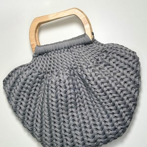 May include: A gray crocheted handbag featuring a thick, textured braided stitch pattern and a light-colored wooden handle. The bag has a rounded, shell-like shape with the gray yarn gathered securely at the top where it attaches to the handle.