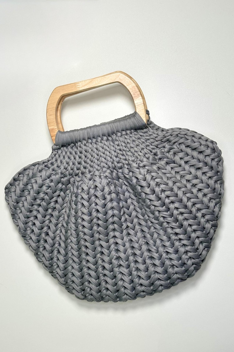 May include: A gray crocheted handbag featuring a thick, textured braided stitch pattern and a light-colored wooden handle. The bag has a rounded, shell-like shape with the gray yarn gathered securely at the top where it attaches to the handle.